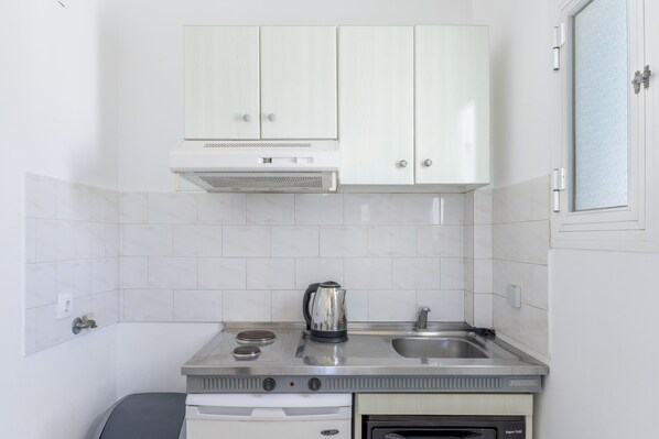 Una cocina compacta con armarios blancos, un lavamanos de acero inoxidable, un hervidor y un horno empotrado.