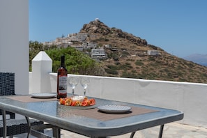 Una mesa con una botella, vasos y platos de comida en un balcón con vistas a un pueblo en la ladera.