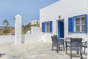 Un bâtiment blanc avec des volets bleus, une terrasse en pierre et des meubles en osier.