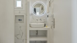 Un baño con pared de mármol, un espejo redondo con luz, un lavabo blanco y una pequeña decoración de peces.