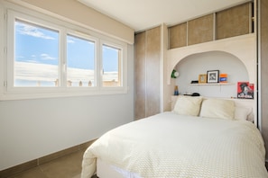 A bedroom with a large bed, a window with a view of the sky, a shelf with books and a camera, and a door with a towel rack. - Bright 1-Bedroom Apartment with Sea View, 10 Minutes from the Beach (Cagnes sur Mer)
