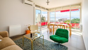 A living room with a green armchair, a glass coffee table, a sofa, a dining table with chairs, and a view of the outdoors through large windows. - Bright 1-Bedroom Apartment with Sea View, 10 Minutes from the Beach (Cagnes sur Mer)