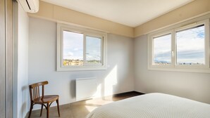 A bedroom with two windows, a radiator, a wooden chair, and a bed with a patterned bedspread. - Bright 1-Bedroom Apartment with Sea View, 10 Minutes from the Beach (Cagnes sur Mer)
