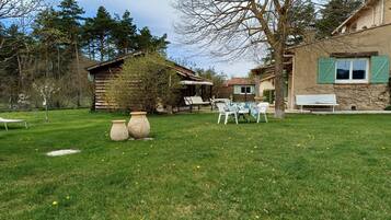 A rustic outdoor area with a wooden cabin, a stone building with green shutters, and a grassy lawn with dandelions.