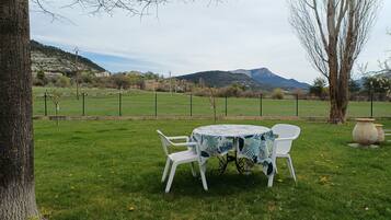 A grassy outdoor area with a table and chairs, a tree, and a view of a mountain range.