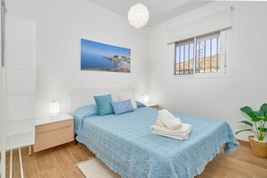 A bedroom with a bed, a nightstand, a bedside lamp, a window with bars, a wall-mounted picture, and a potted plant.