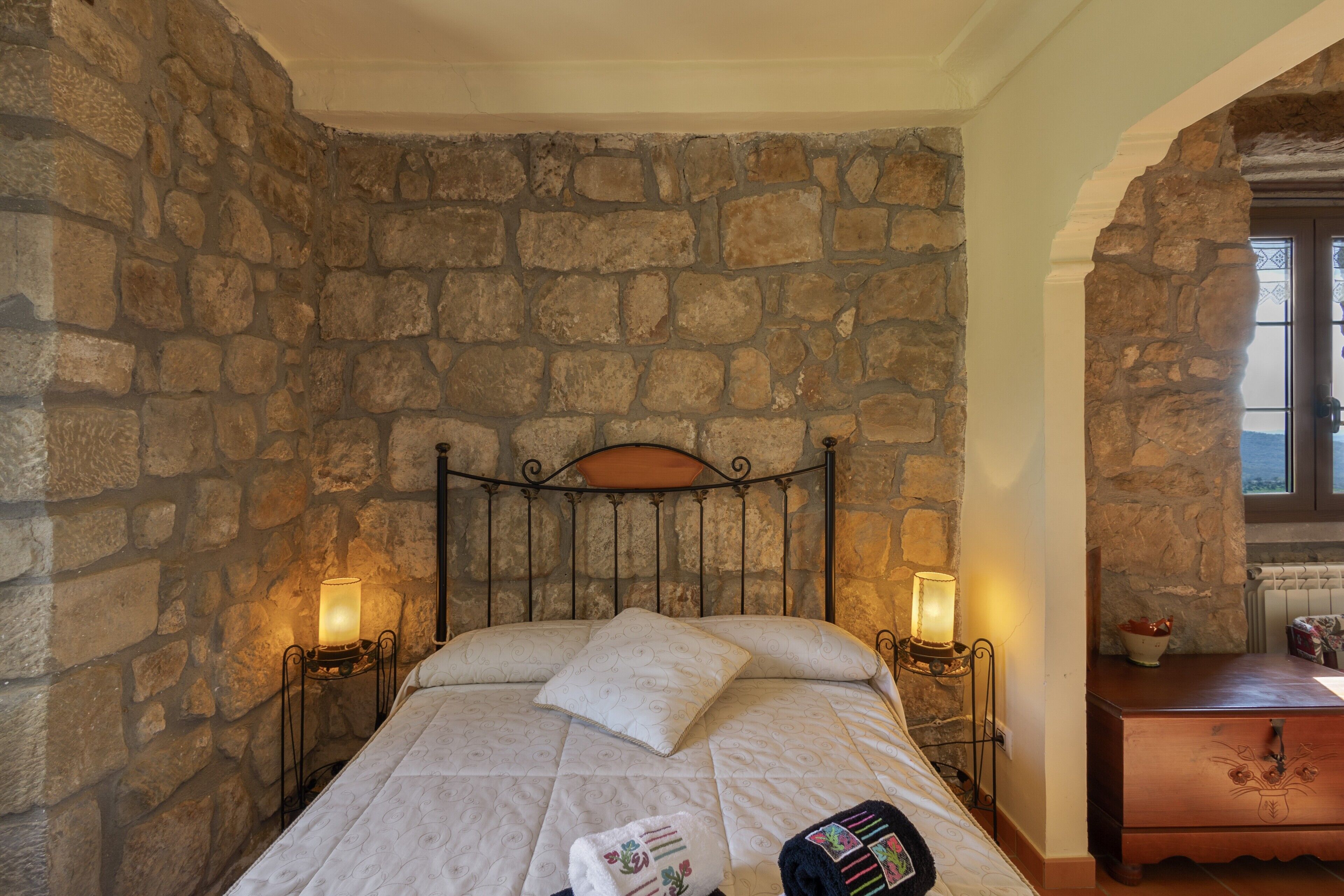 A bedroom with a stone wall, a bed with a headboard, two bedside lamps, and a nightstand with a lamp.