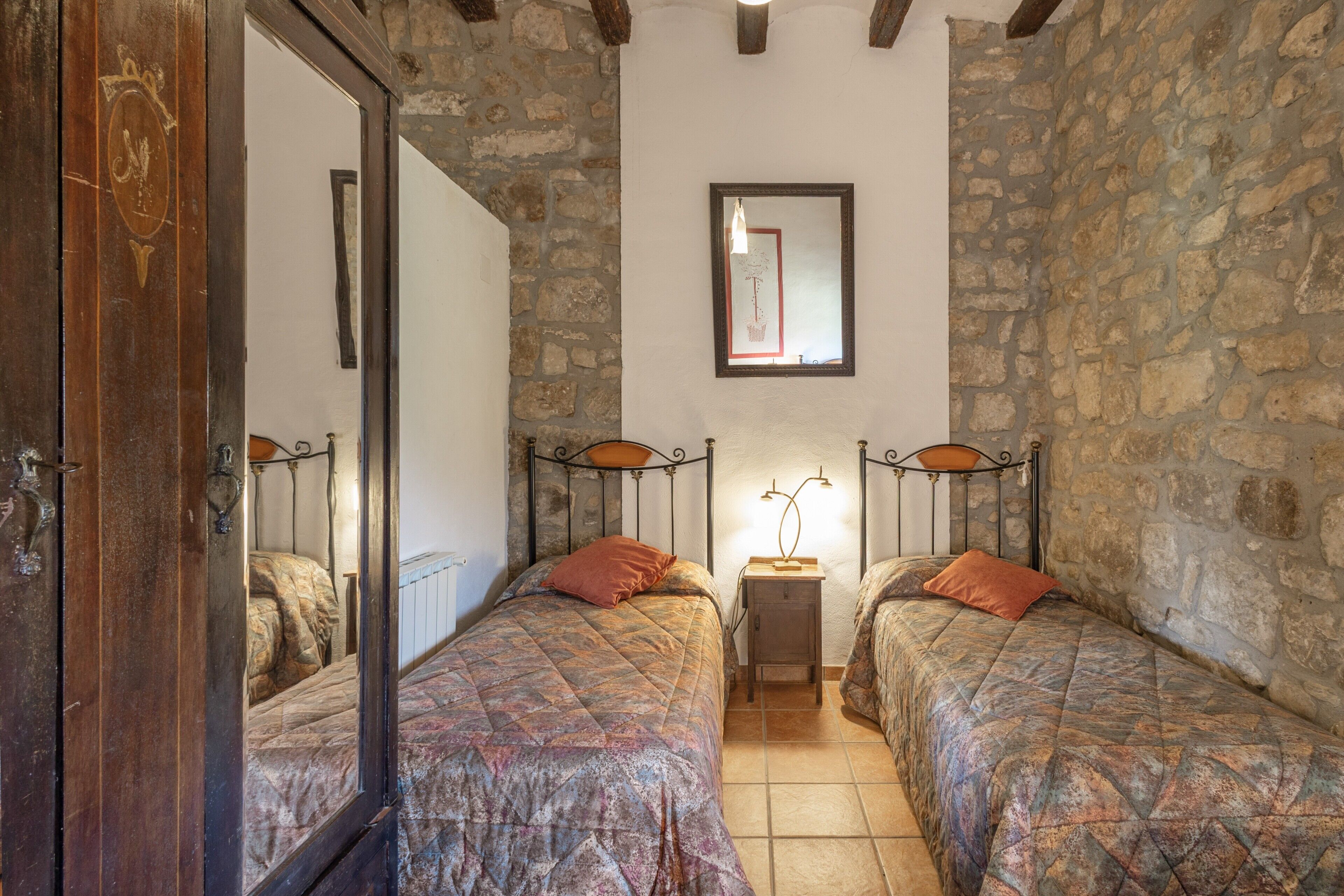 A room with two beds, a stone wall, a mirror, and a small table with a lamp.
