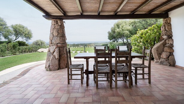 Une terrasse extérieure agrémentée d’une colonne en pierre, d’une table en bois et de chaises, offrant une vue sur un jardin aménagé et un paysage lointain.