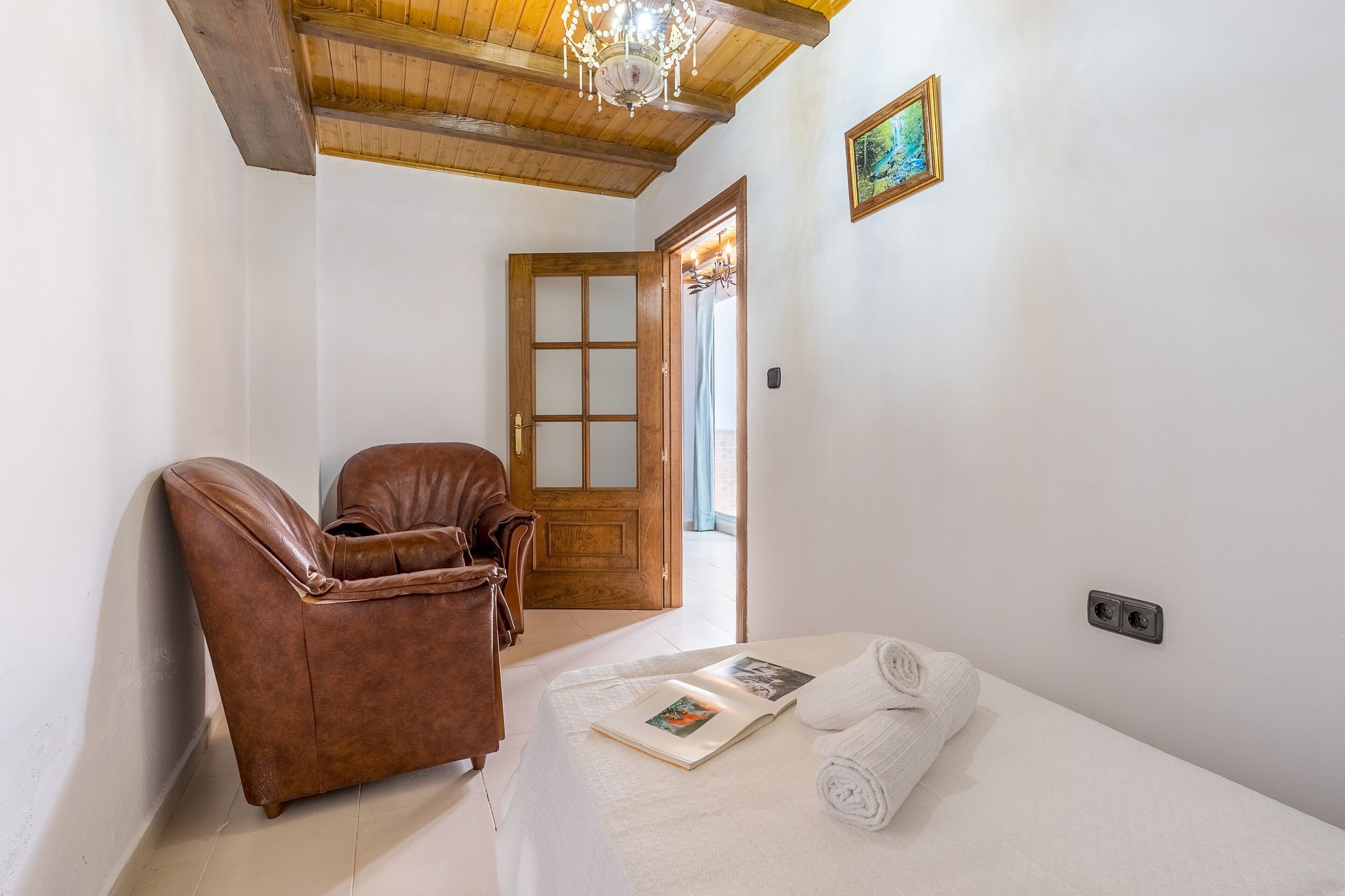 Una habitación con techo de madera, un sillón de cuero marrón, una cama con manta blanca y una puerta que da a otra habitación.