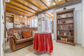 A cozy living area with a leather sofa, a red table, wooden shelves with decorative items, and a kitchen in the background.