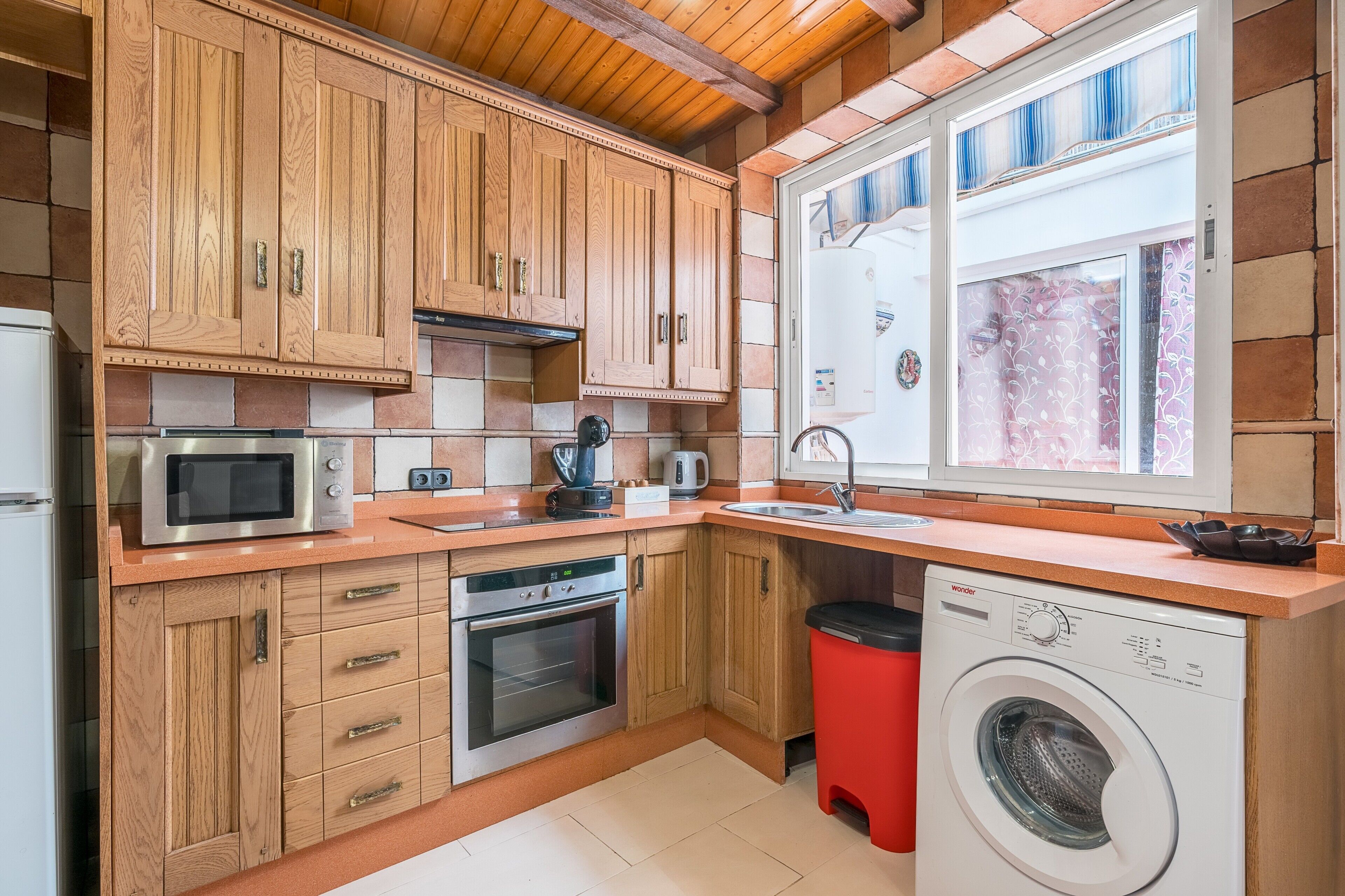 Cocina compacta con armarios de madera, microondas, horno, lavaplatos y lavadora.