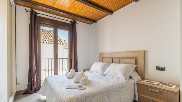 Ein Schlafzimmer mit einem Bett, einem Nachttisch, einem Holz-Kopfteil, einem Fenster mit Vorhängen und einem Balkon.