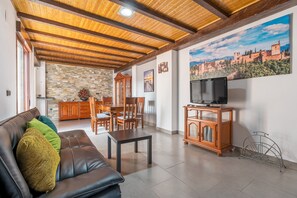 Un salon avec un plafond en bois, un mur en pierre, un coin repas et un téléviseur posé sur un meuble en bois.