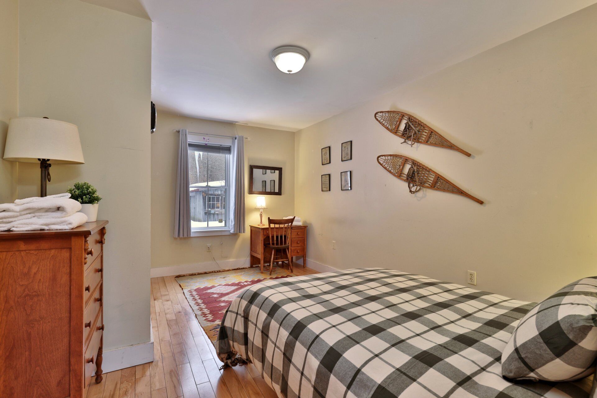 Ein Schlafzimmer mit einem Holzkapuzen, einem Bett mit kariertem Überwurf, einem Stuhl, einem Schreibtisch, einem Fenster mit Vorhängen und Wanddekorationen.