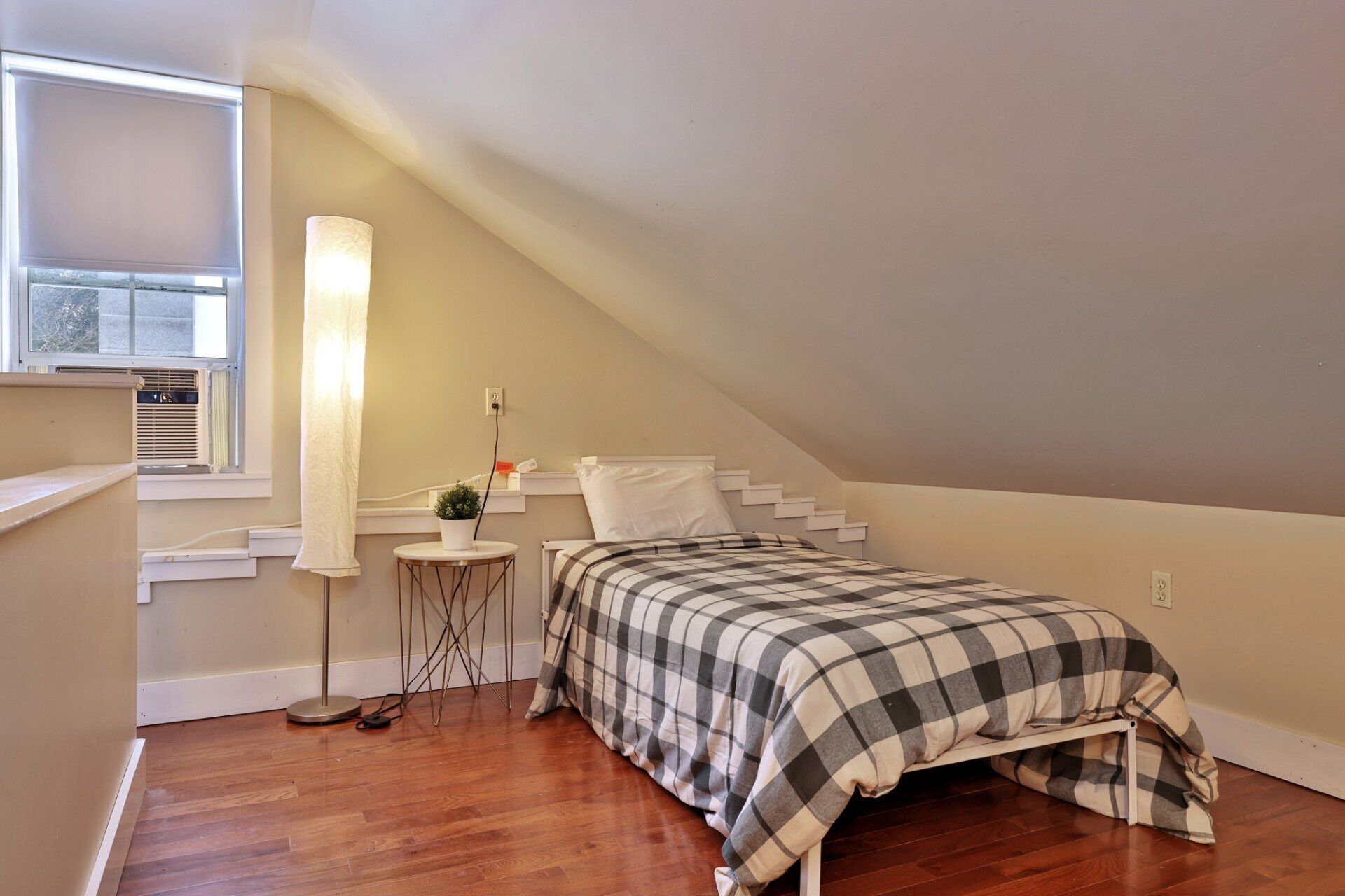 Une petite chambre mansardée, chaleureuse et accueillante, avec un plafond en pente, un lit simple recouvert d’une courtepointe à carreaux, une table de chevet avec une lampe et une fenêtre équipée de stores.