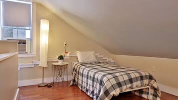Une petite chambre mansardée, chaleureuse et accueillante, avec un plafond en pente, un lit simple recouvert d’une courtepointe à carreaux, une table de chevet avec une lampe et une fenêtre équipée de stores.