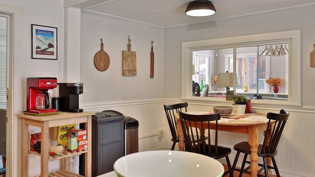 A dining area with a wooden table, four chairs, a coffee maker, and a trash can. - Chittenden Farmhouse: Historic 4BR/2BA, 20 Min to Killington, 7 Min to Mountaintop Resort (Chittenden)