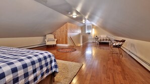 A spacious attic bedroom with a large bed, a wooden chair, a small table, and a staircase leading to an upper level. - Chittenden Farmhouse: Historic 4BR/2BA, 20 Min to Killington, 7 Min to Mountaintop Resort (Chittenden)