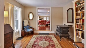 A cozy living room with a wooden floor, a red and beige rug, a blue armchair, a wooden dresser, and a bookshelf filled with books. - Chittenden Farmhouse: Historic 4BR/2BA, 20 Min to Killington, 7 Min to Mountaintop Resort (Chittenden)