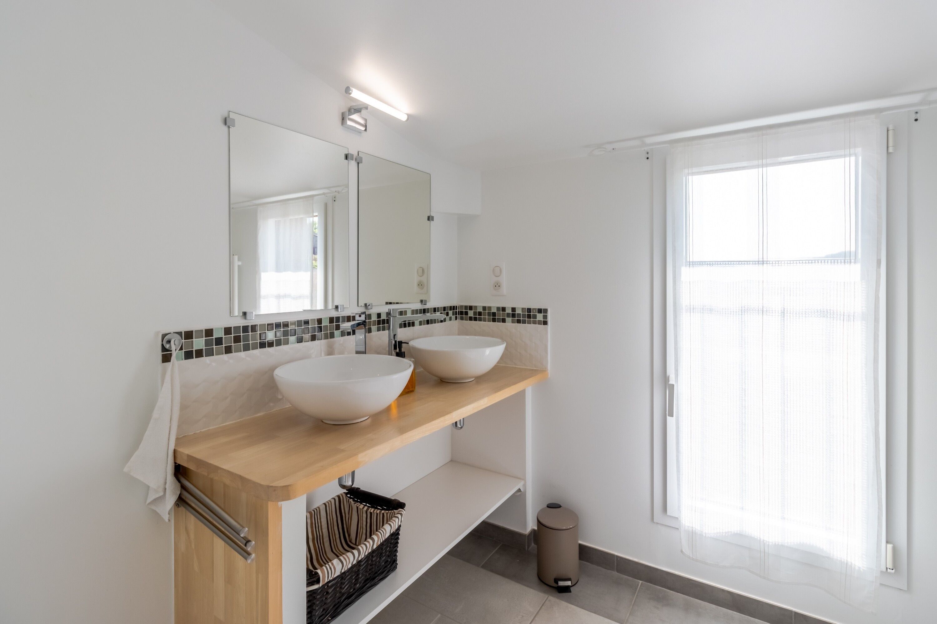Un baño moderno con dos lavamanos grandes, un espejo y una ventana con cortinas translúcidas.