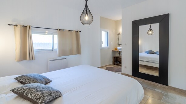 A bedroom with a bed, two pillows, a hanging light fixture, a window with curtains, a mirror, and a bathroom visible through an open door. - Le Séchoir – Authentic House with Private Pool in Périgord Noir, Perfect for Families or Groups (Coly-Saint-Amand)