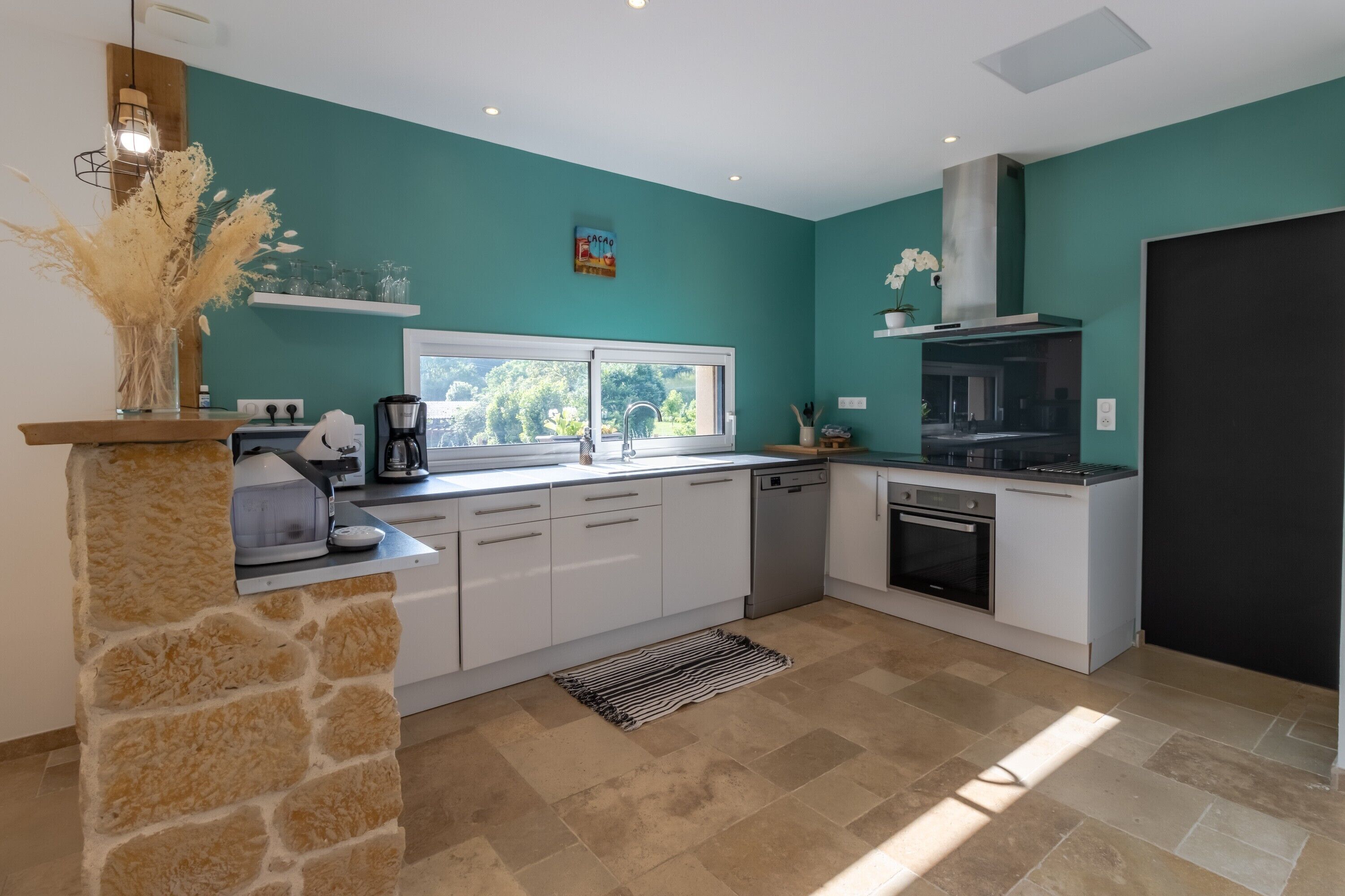 A modern kitchen with white cabinets, a stone accent wall, a built-in oven, and a window providing natural light.
