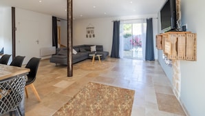 A modern living room with a sofa, dining table, and chairs, a TV mounted on a wall, and a large window with curtains. - Le Séchoir – Authentic House with Private Pool in Périgord Noir, Perfect for Families or Groups (Coly-Saint-Amand)