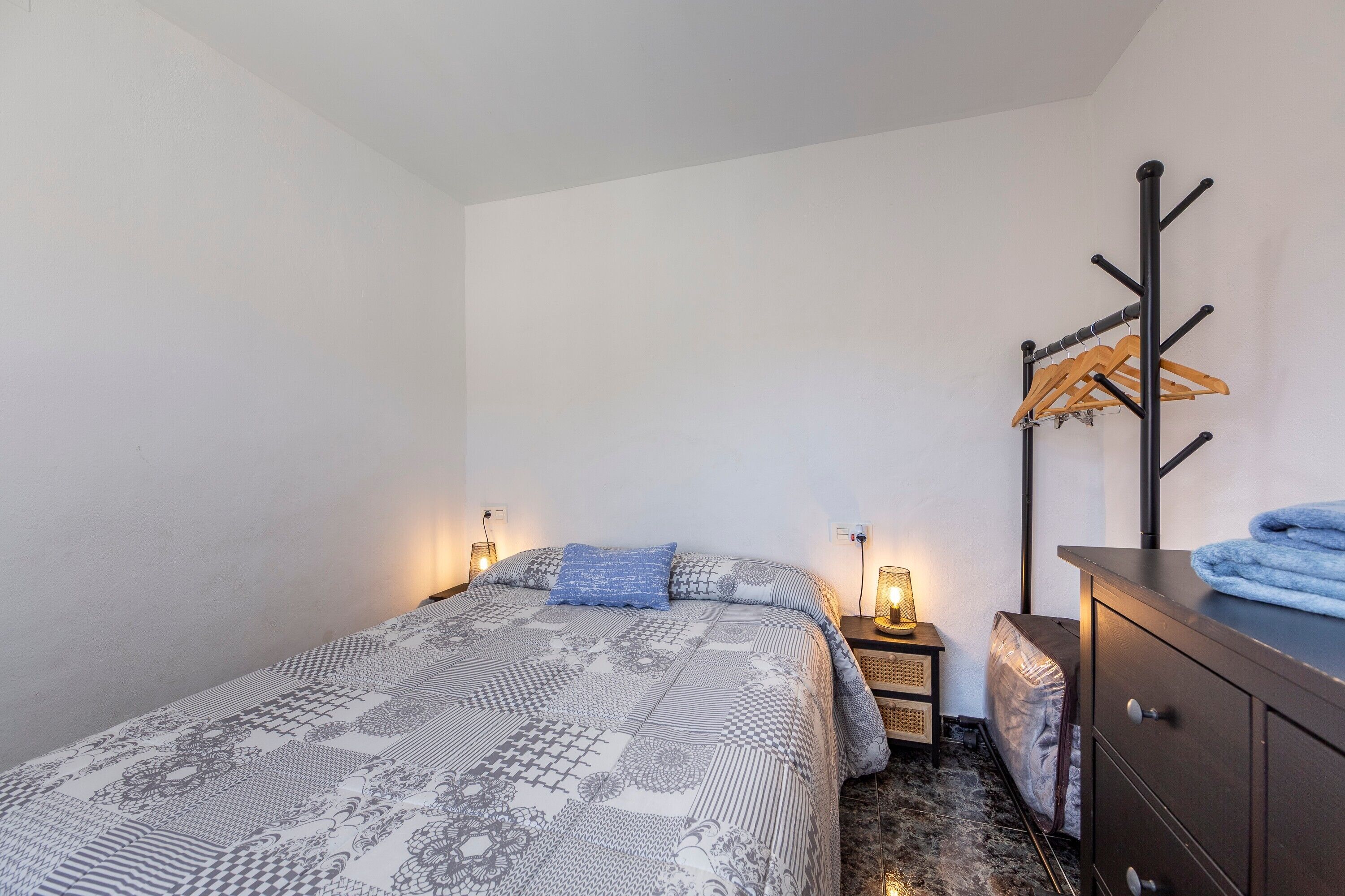 A bedroom with a patterned bedspread, a bedside table with a lamp, a wooden coat rack, and a dresser with a towel on top.