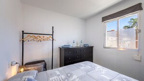 A bedroom with a bed, a dresser, a window with blinds, a hanging rack, and a bedside lamp.