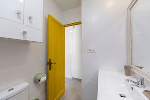 A bathroom with white tiles, a yellow door, a toilet, a sink with a mirror, and a trash bin.