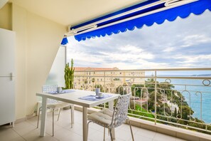 Un balcón con mesa y sillas, vista al mar y edificios al fondo.