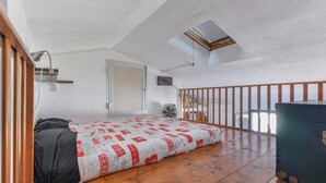 Eine kleine Einzimmerwohnung mit Schlafgalerie, einem Bett, einem Ventilator, einem Koffer und Blick ins Freie.