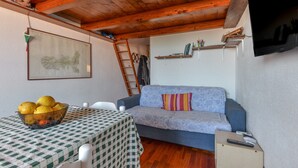 Ein Zimmer mit Holzboden, einer Couch, einem Tisch mit kariertem Tischläufer, einer Obstschale und einem an der Wand befestigten Fernseher.