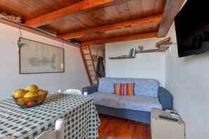 A room with a wooden ceiling, a sofa, a table with a checkered tablecloth, a bowl of fruit, and a mounted TV.