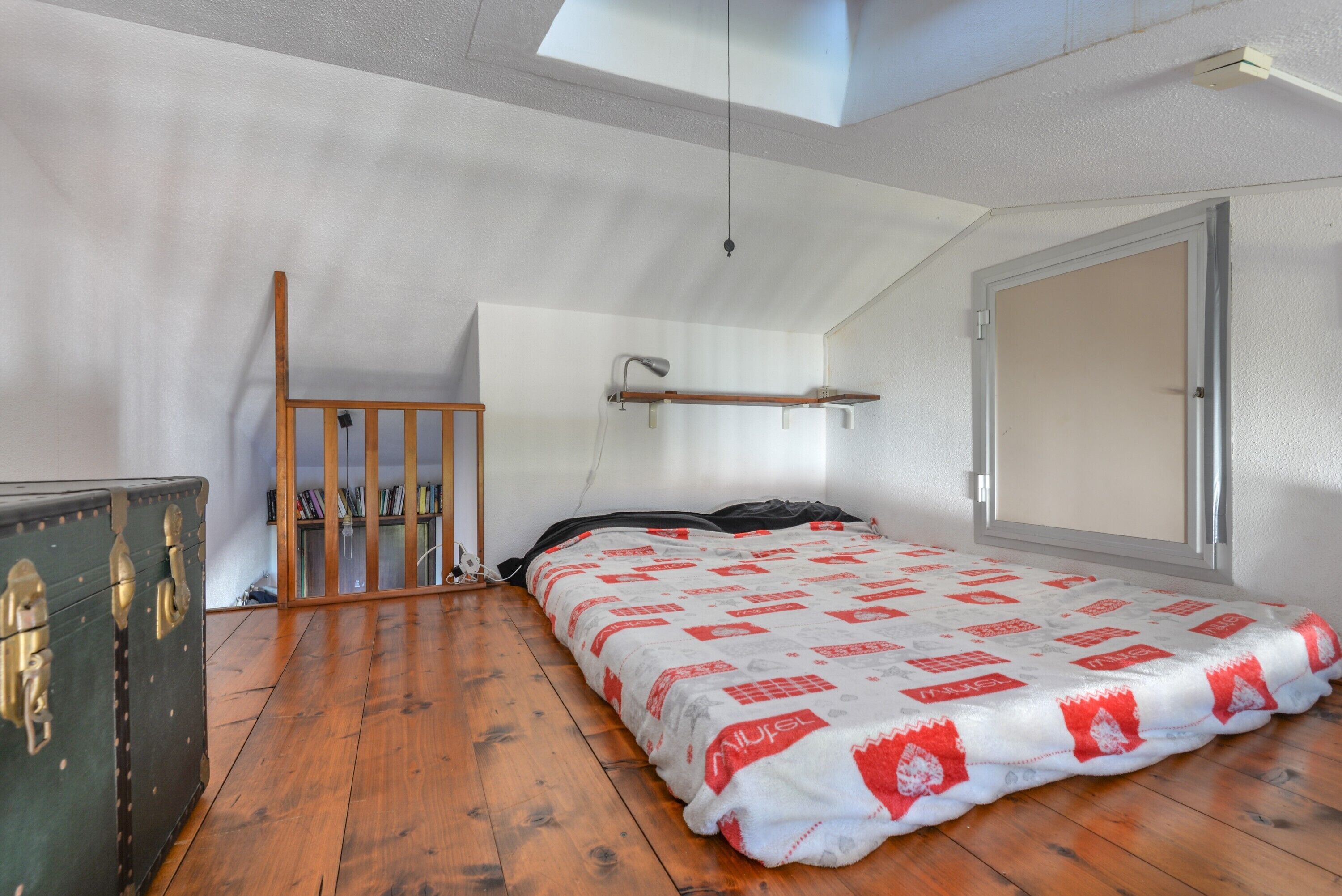 A bedroom with a sloped ceiling, a bed with a patterned blanket, a wooden dresser, a desk with a lamp, and a window.