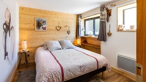 Une chambre à coucher avec une tête de lit en bois, un lit recouvert d’une couvre-lit à motifs en forme de cœur, une lampe de chevet, un radiateur fixé au mur et une fenêtre donnant sur un paysage agréable.