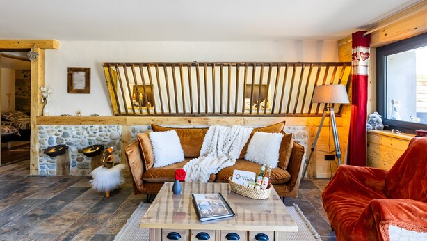 Une pièce chaleureuse avec un mur lambrissé en bois, un canapé marron avec des coussins blancs, une table basse avec un livre et un panier, et un fauteuil rouge.