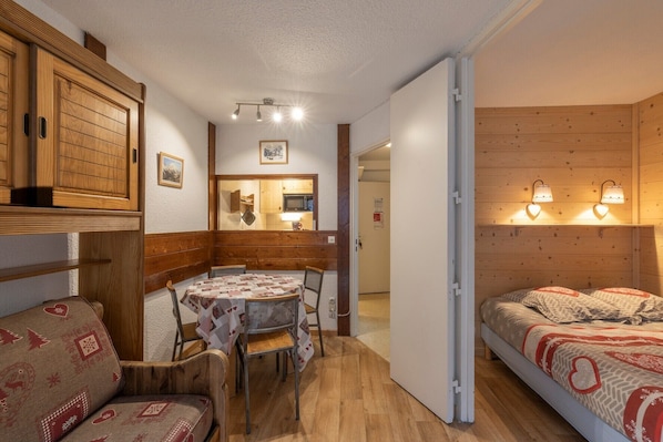 A cozy room with a wooden bunk bed, a small dining table with chairs, a kitchen area, and a hallway leading to another room.