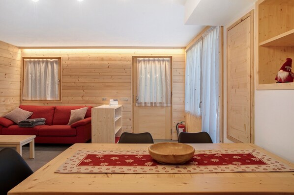 A cozy room with a wooden interior, a red sofa, a dining table with a wooden bowl, and a small shelf with a figurine.