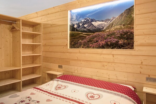 A wooden room with a bed, a wooden wardrobe, and a wall-mounted picture of a mountain landscape.