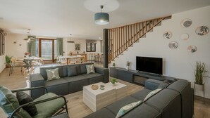 Ein modernes Wohnzimmer mit einer Couch, einem Couchtisch und einem Fernseher. Im Hintergrund ist ein Essbereich mit einer Treppe und Holzhandläufen zu sehen.