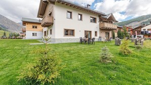 A two-story building with a balcony, a grassy yard, and mountains in the background. - Studio 'Pino Mugo' with Mountain View, Balcony and Wi-Fi (Livigno)