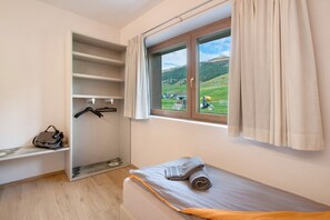 Een slaapkamer met een bed, een raam met gordijnen, een kledingkast en een klein bankje met een handtas.