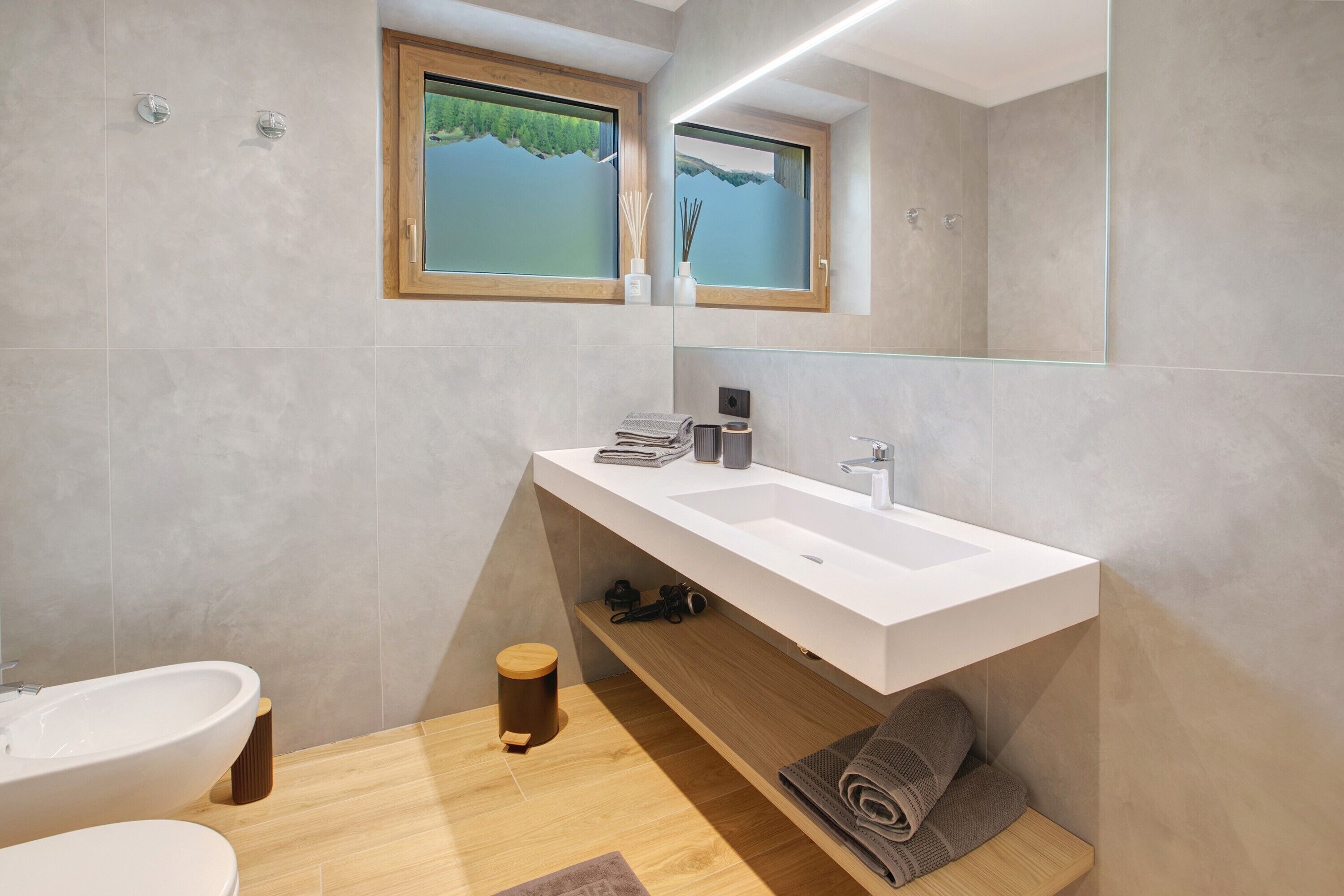 Un bagno moderno con un lavandino capiente, un water, un bidet, un mobile bagno in legno e una finestra vista lago.