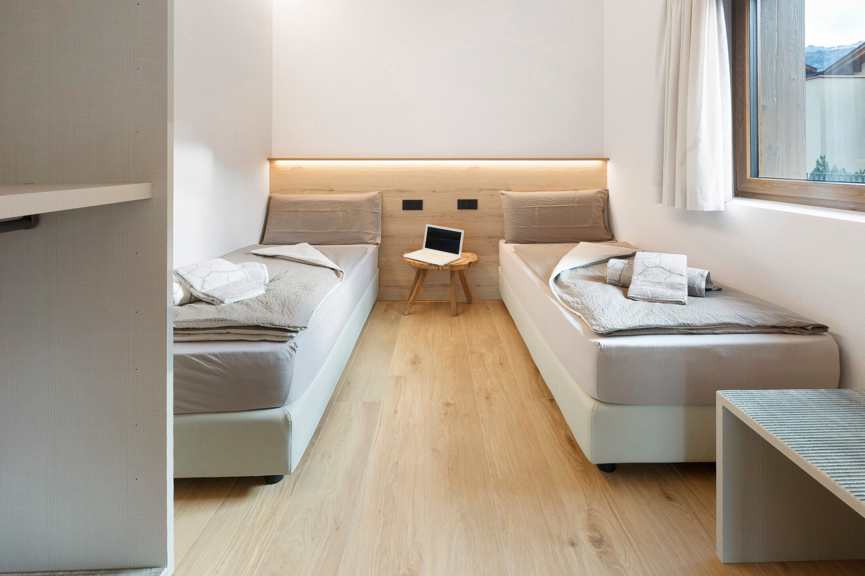 A compact room with two beds, a small table with a laptop, and a window with curtains.