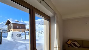 A room with a large sliding glass door leading to a snowy outdoor area, a wooden floor, and a sofa with cushions. - Apartment 'Stella Alpina' with Mountain View, Private Garden and Wi-Fi (Livigno)