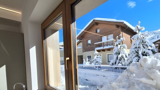 A window with a view of a building covered in snow, a wooden balcony, and a white sink with a blue cloth. - Apartment 'Stella Alpina' with Mountain View, Private Garden and Wi-Fi (Livigno)