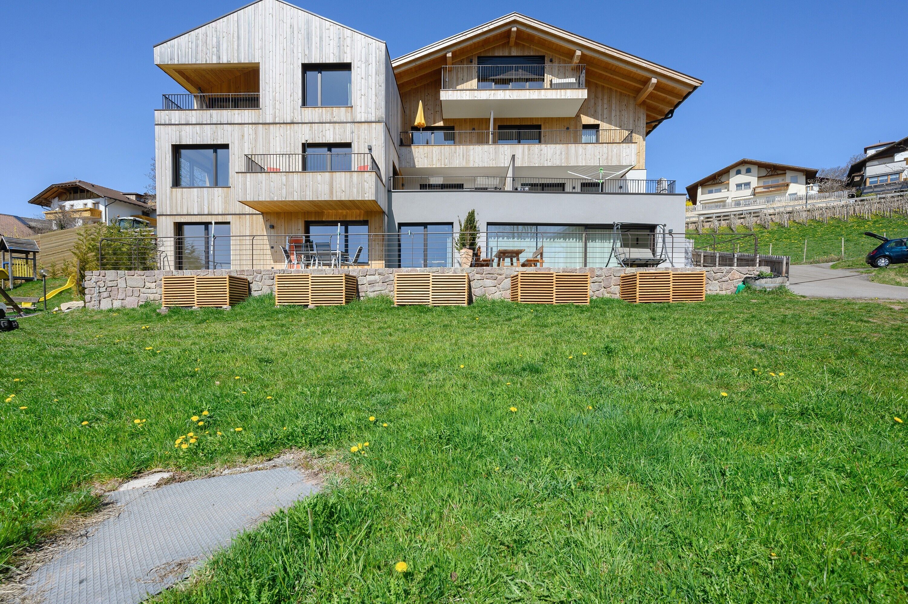 A modern multi-story building with balconies, a stone retaining wall, and a grassy area in front.