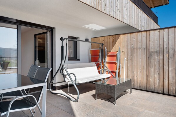 Un espace extérieur moderne, aménagé avec une table en verre, des chaises en métal, un banc blanc rembourré et un fauteuil en osier.